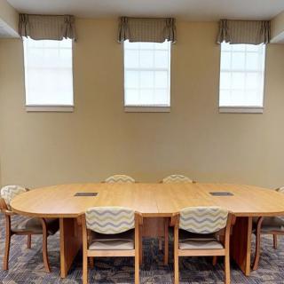 A conference room with a large wooden table and several chairs, positioned below three windows.