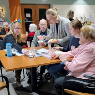 A group of elderly individuals enjoying a social activity at a community center, with a caregiver assisting them.