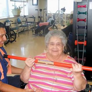 An elderly woman participates in an exercise session with assistance from a caregiver in a healthcare facility.