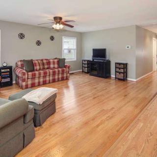 A spacious living room with hardwood flooring, featuring a plaid sofa, armchair, ceiling fan, and a television set.