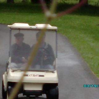 Two people driving a golf cart on a paved path surrounded by grass.