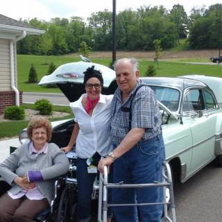Three people posing outside with a classic car, including an elderly couple using mobility aids.