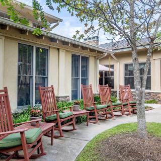 A row of wooden rocking chairs with green cushions outside a building surrounded by a garden.