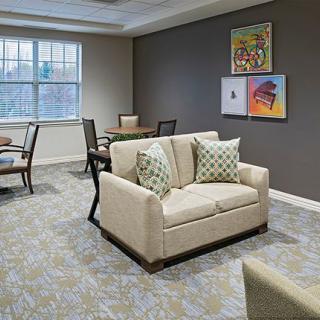 A cozy living room featuring a beige sofa with patterned cushions, wall art, and ample natural light from a large window.