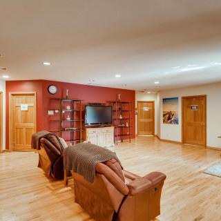 A cozy living room with wooden flooring, featuring comfortable chairs and a television on a red accent wall.
