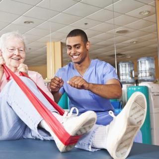 An elderly woman is assisted by a healthcare professional during a physical therapy session using an exercise band.