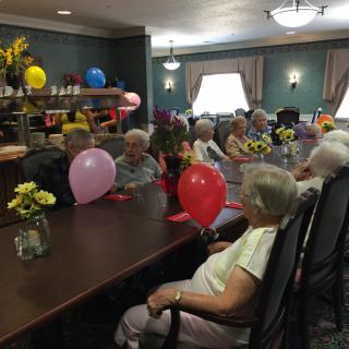 A group of seniors enjoying a celebration with balloons and flowers in an indoor setting.
