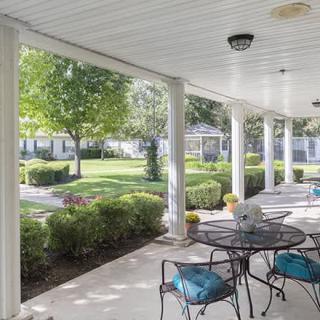 A covered patio with outdoor furniture overlooks a well-manicured garden featuring trees and shrubs.