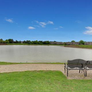 A peaceful view of a lake with a bench on the grassy shore under a clear blue sky.