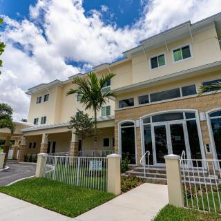 A modern residential building with large windows and a small front yard, set against a partly cloudy sky.