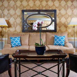 A cozy living room interior with a central coffee table adorned with an orchid plant, a beige sofa, and blue patterned throw pillows, accented by two floor lamps and a decorative mirror on the wall.