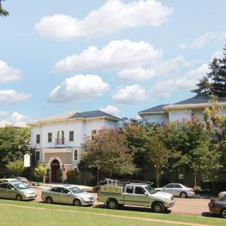A view of a stately building with a row of cars parked in front, surrounded by trees against a backdrop of blue sky and clouds.