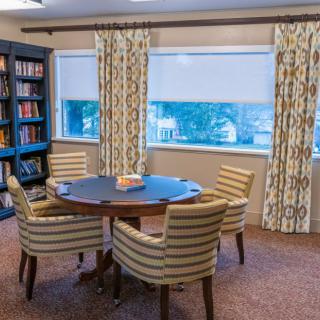 A cozy reading room with a round table and striped chairs, surrounded by bookshelves and windows.