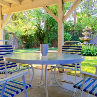 A sunny patio with a table and striped chairs, overlooking a lush garden and fountain.