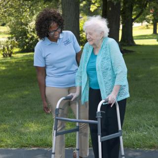 A caregiver assists an elderly woman using a walker outdoors.