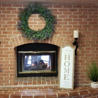 A cozy brick fireplace decorated with a wreath, home sign, candle, and indoor plant.