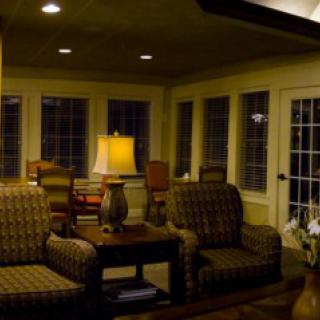 A cozy living room featuring patterned chairs, a table with a lamp, and large windows.