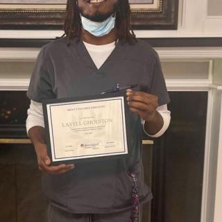 A person wearing scrubs and a mask holds up a certificate indoors.