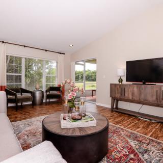 A cozy living room featuring a sofa, coffee table, TV, and large windows letting in natural light.