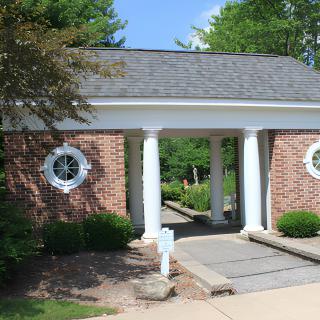 A small brick building with white columns and circular windows, surrounded by greenery.