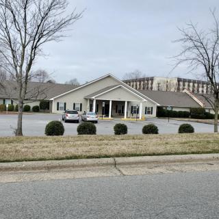 A building with a parking lot and several cars, surrounded by trees and bushes on an overcast day.