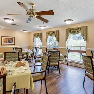 A cozy dining room with neatly arranged tables and chairs, featuring large windows with draped curtains and a ceiling fan.
