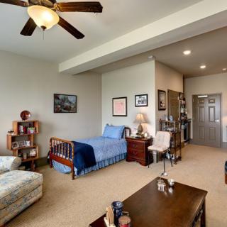 A cozy bedroom featuring a single bed, bookshelf, and armchair with a ceiling fan overhead.