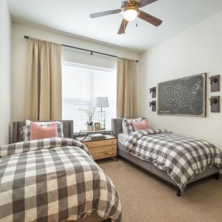 A cozy bedroom with twin beds featuring checkered bedding and decorative pillows, complemented by a nightstand and lamp beneath large curtains and a ceiling fan.