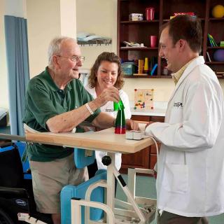 An elderly man receives assistance from a medical professional and caregiver during a physical therapy session.