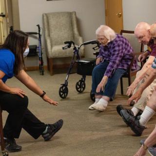 A group of seniors participating in a stretching exercise led by an instructor in a care facility.