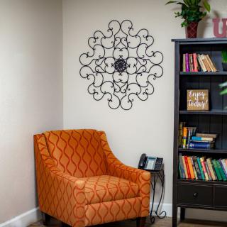 A cozy reading nook with an orange patterned chair, a decorative bookshelf filled with books, and intricate wall art.