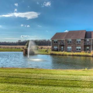 A serene pond with a fountain alongside a brick building under a clear blue sky.