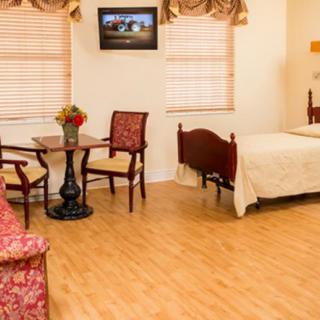 A cozy bedroom with classic furniture, including a bed, chairs, and a small table, featuring wood flooring and a mounted television.
