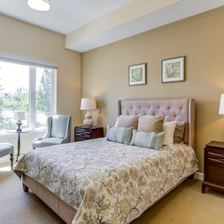A cozy bedroom featuring a tufted headboard, elegant bedding, and two nightstands with lamps. A large window allows natural light to illuminate the room.