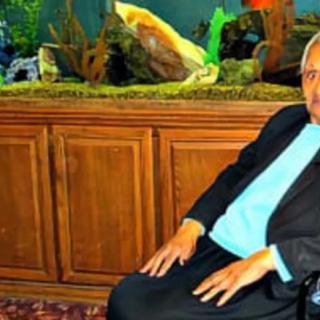 A man in a suit sitting beside an aquarium indoors.