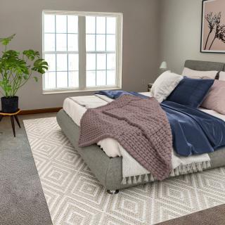 A modern bedroom featuring a neatly-made bed with pillows and throws, a large window, a house plant, and a patterned rug.