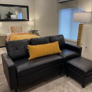 A cozy living room with a black sofa and ottoman, complemented by a yellow pillow, near a bed with warm lighting.