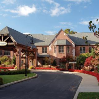 A scenic view of a brick building with a landscaped entrance and driveway under a partly cloudy sky.