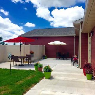 A sunny backyard patio with outdoor furniture and umbrellas, surrounded by a grassy area and a fence.