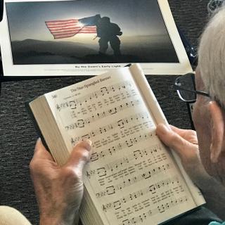 An elderly person reading a music sheet, with a photograph of a soldier holding an American flag in the background.