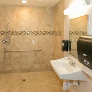 A spacious and accessible bathroom with tiled walls, featuring a sink, mirror, handrails, soap dispenser, and paper towel dispenser.