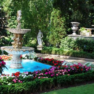 A serene garden scene featuring a central fountain surrounded by vibrant flowers and lush greenery, with a classical statue in the background.