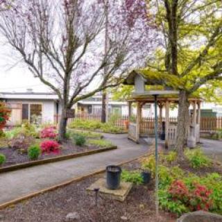 A serene garden with a gazebo, surrounded by trees and well-maintained flower beds, featuring a winding pathway.