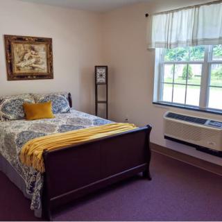 A cozy bedroom featuring a decorated bed with a patterned quilt and pillows, an air conditioner under the window, and framed artwork on the wall.