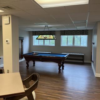 A game room featuring a pool table under a decorative light, with large windows and wooden flooring.