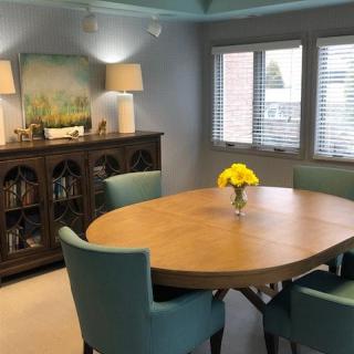 A cozy dining room with a wooden oval table surrounded by teal chairs, featuring a sideboard with lamps and landscape artwork. A vase of yellow flowers adds a touch of color.