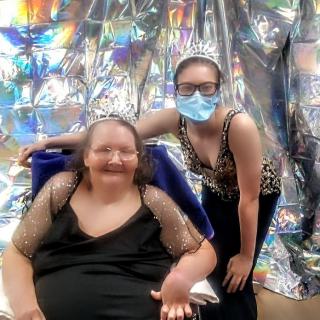 Two people at a party; one is seated in a wheelchair, and the other is standing beside them, wearing a mask and glasses.