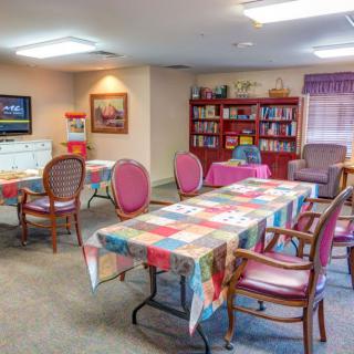 A cozy community room with tables covered in colorful tablecloths, surrounded by chairs. A bookshelf and a television are visible in the background.