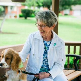 An elderly woman sitting on a bench outdoors, smiling as she pets a dog.