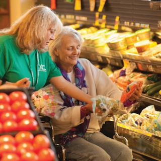 A volunteer assists an elderly woman in selecting fresh produce at a grocery store.